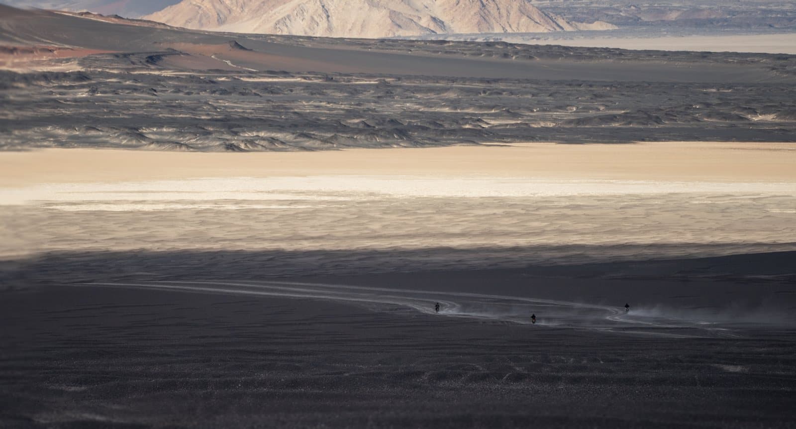 Three motorcycles crossing a high plain between salt flats, black sand, and mountains.